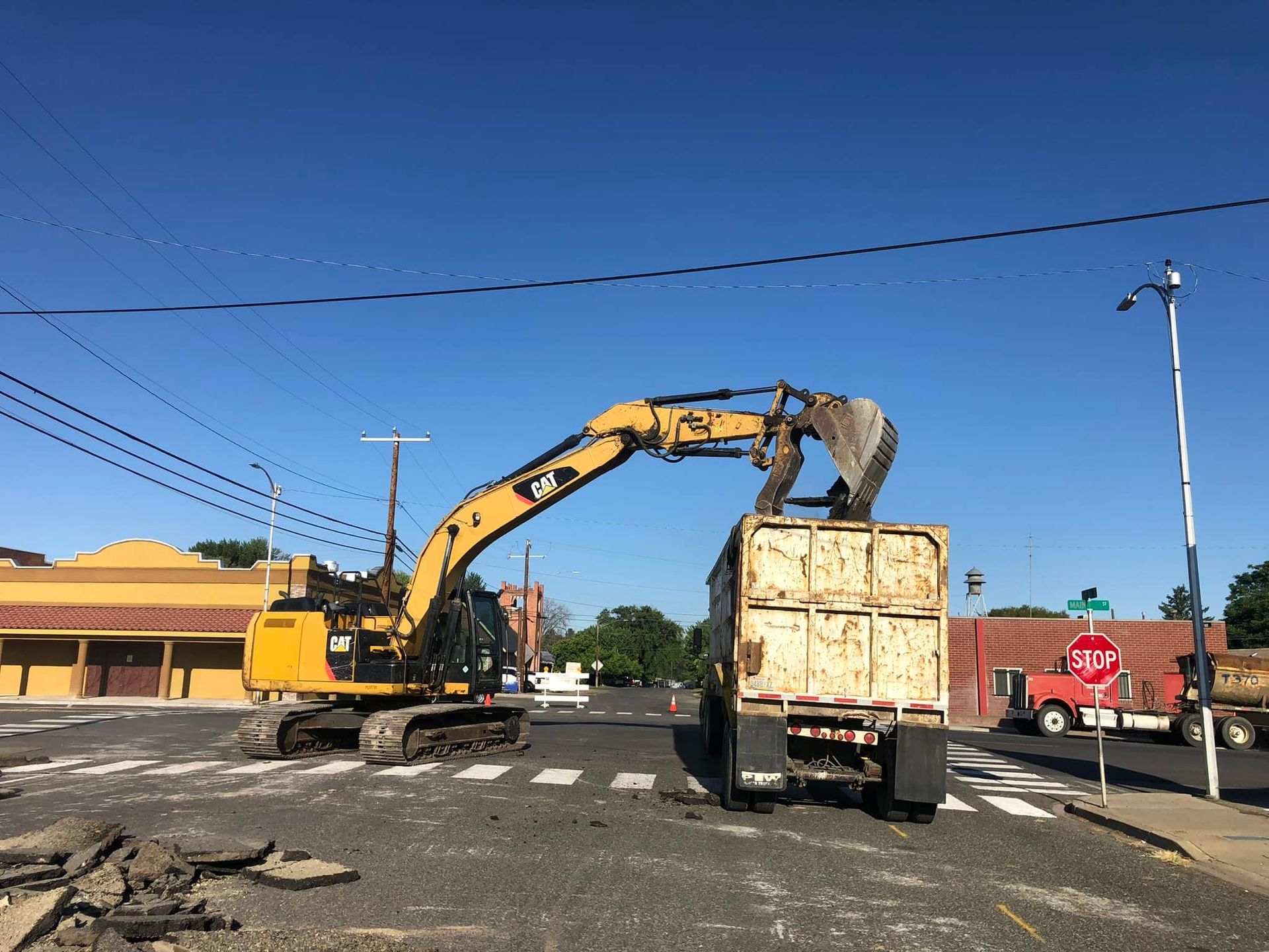 Excavator loading a truck with asphalt debris on a street intersection under a blue sky.