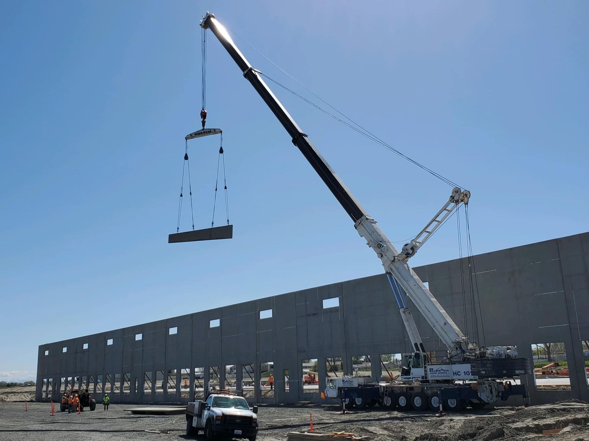 A crane lifts a concrete wall section at a construction site on a sunny day.