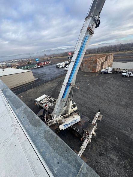 Crane on a rooftop with extended arm, reaching over a gravel lot with trucks and buildings, cloudy sky.