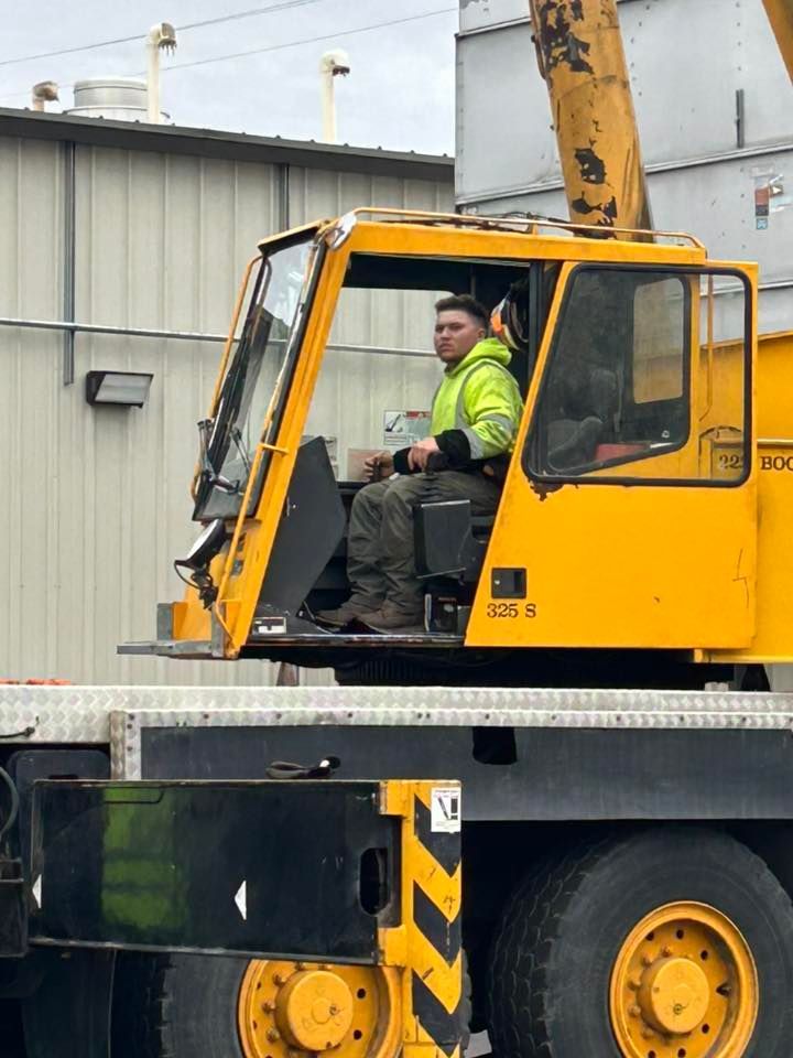 Crane operator in a yellow crane cab, wearing a safety vest, looking toward the camera.