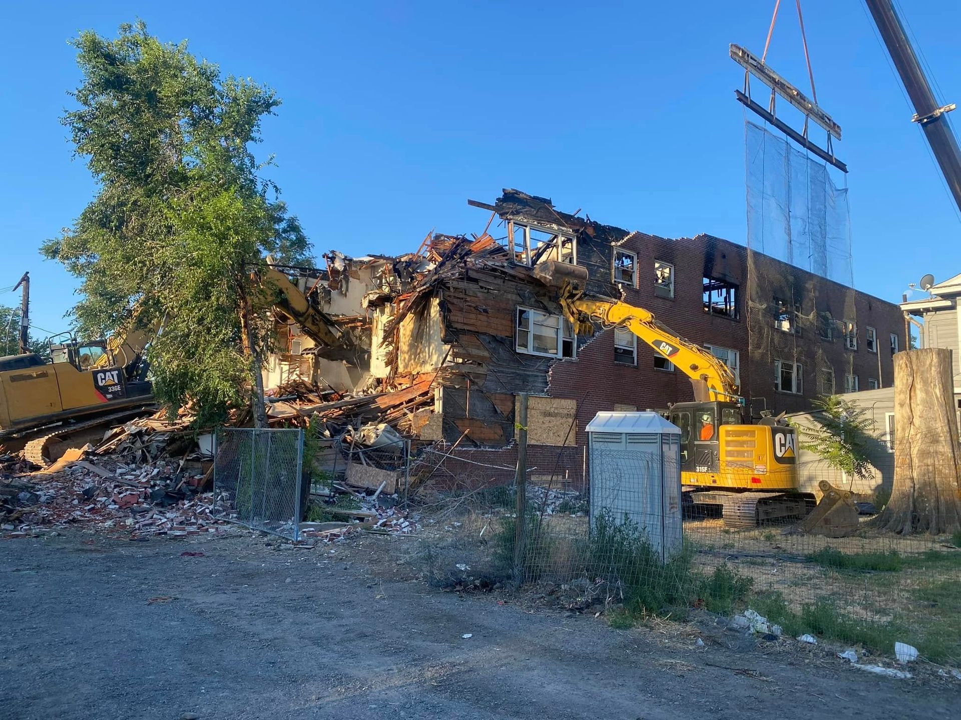 Demolition of a two-story brick building. Yellow excavator tears at the roof, crane with netting present. Debris scattered. Clear sky.