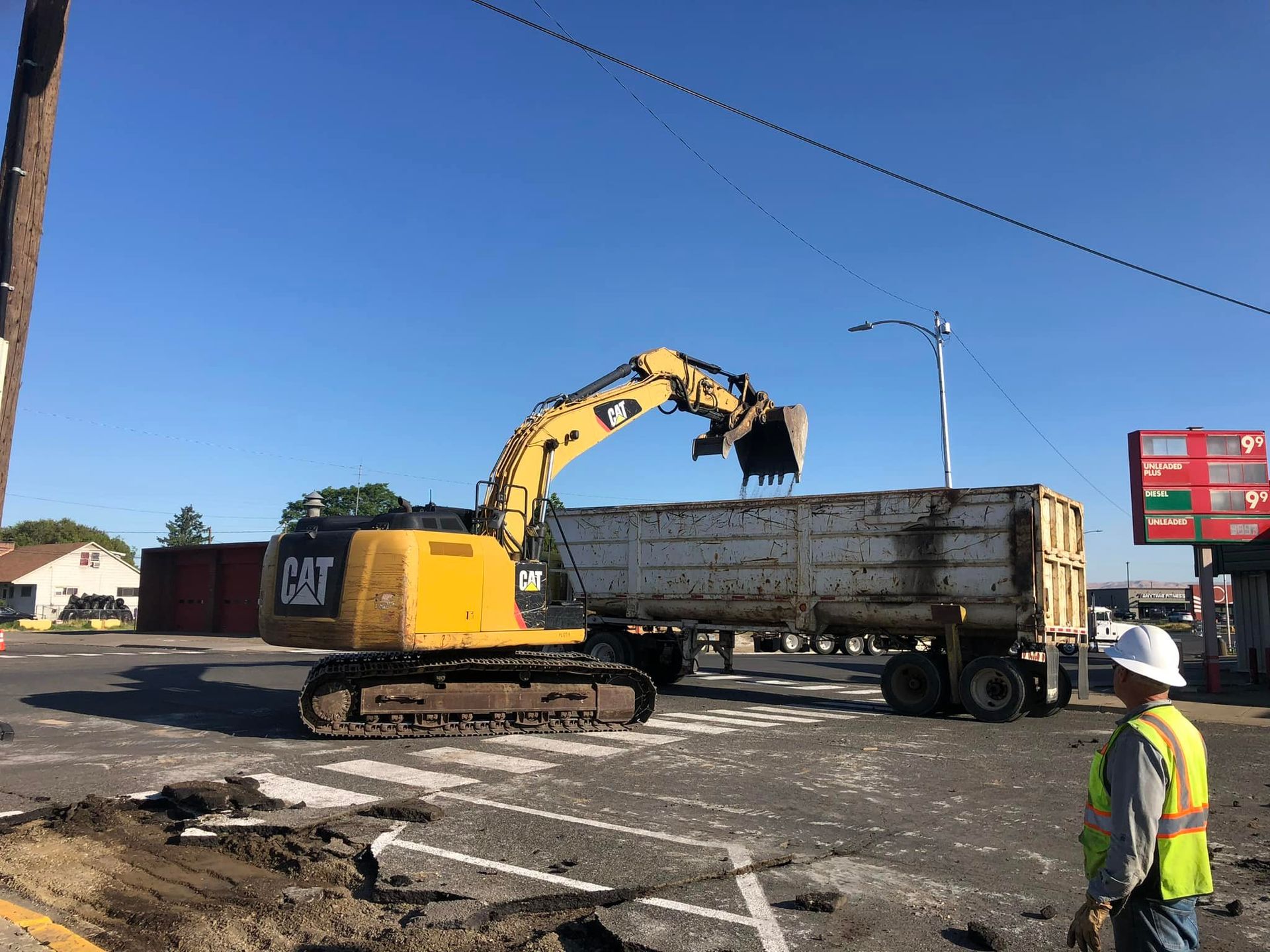A yellow CAT excavator loads debris into a dump trailer at a road construction site. A worker watches.