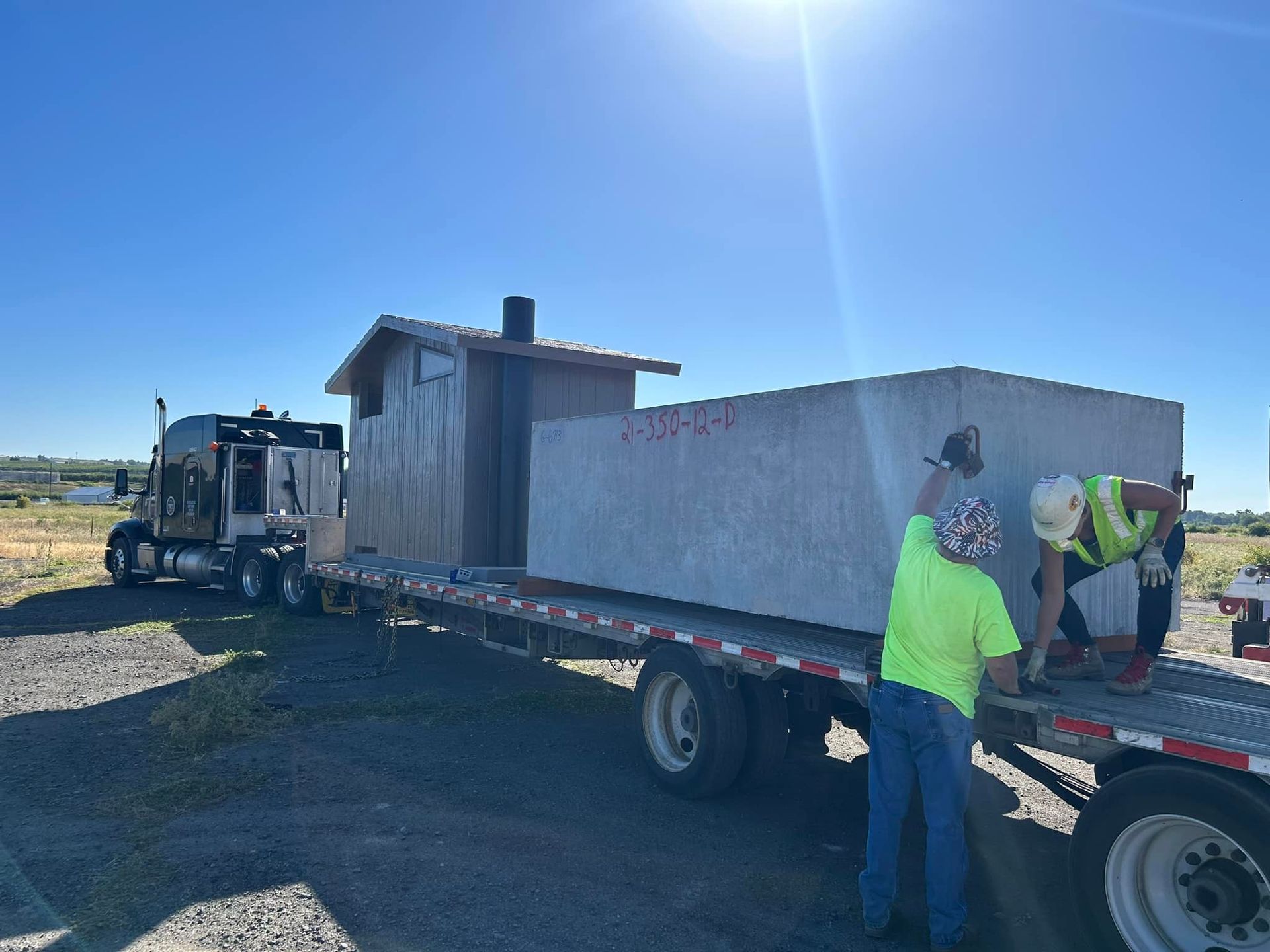Truck hauling large concrete block with two workers securing it on a flatbed trailer under a clear blue sky.
