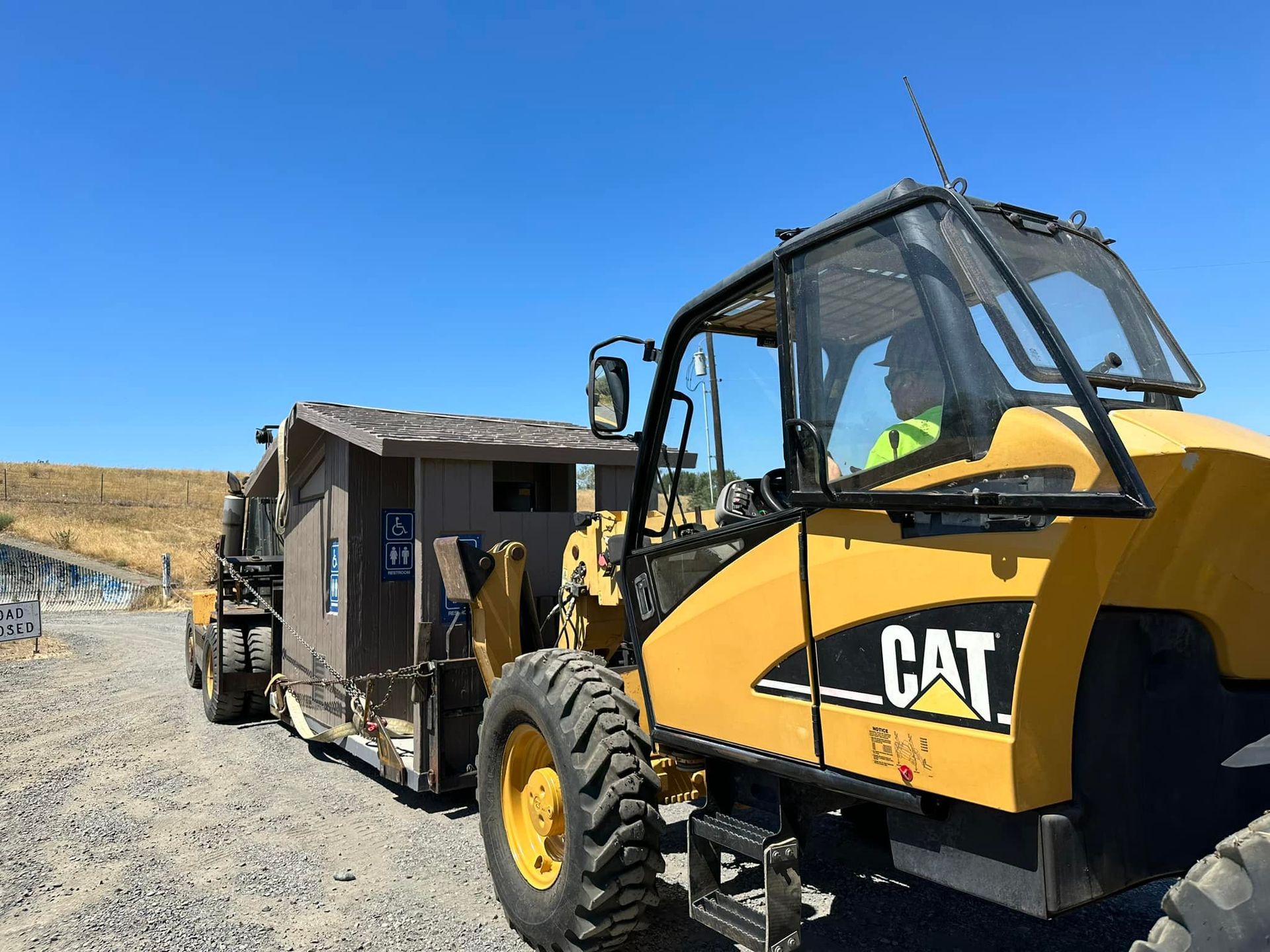 Yellow CAT forklift towing a brown outhouse on a gravel road under a blue sky.