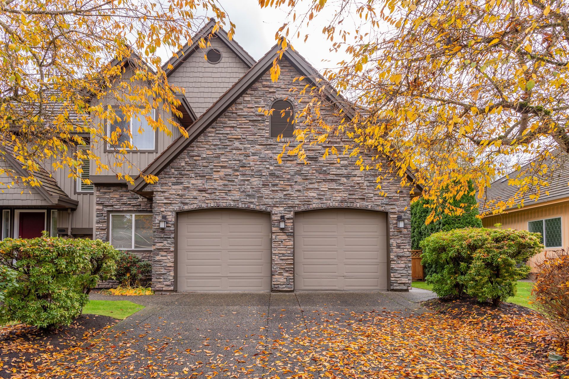 Stone suburban house with two garages under autumn trees and fallen leaves in the driveway