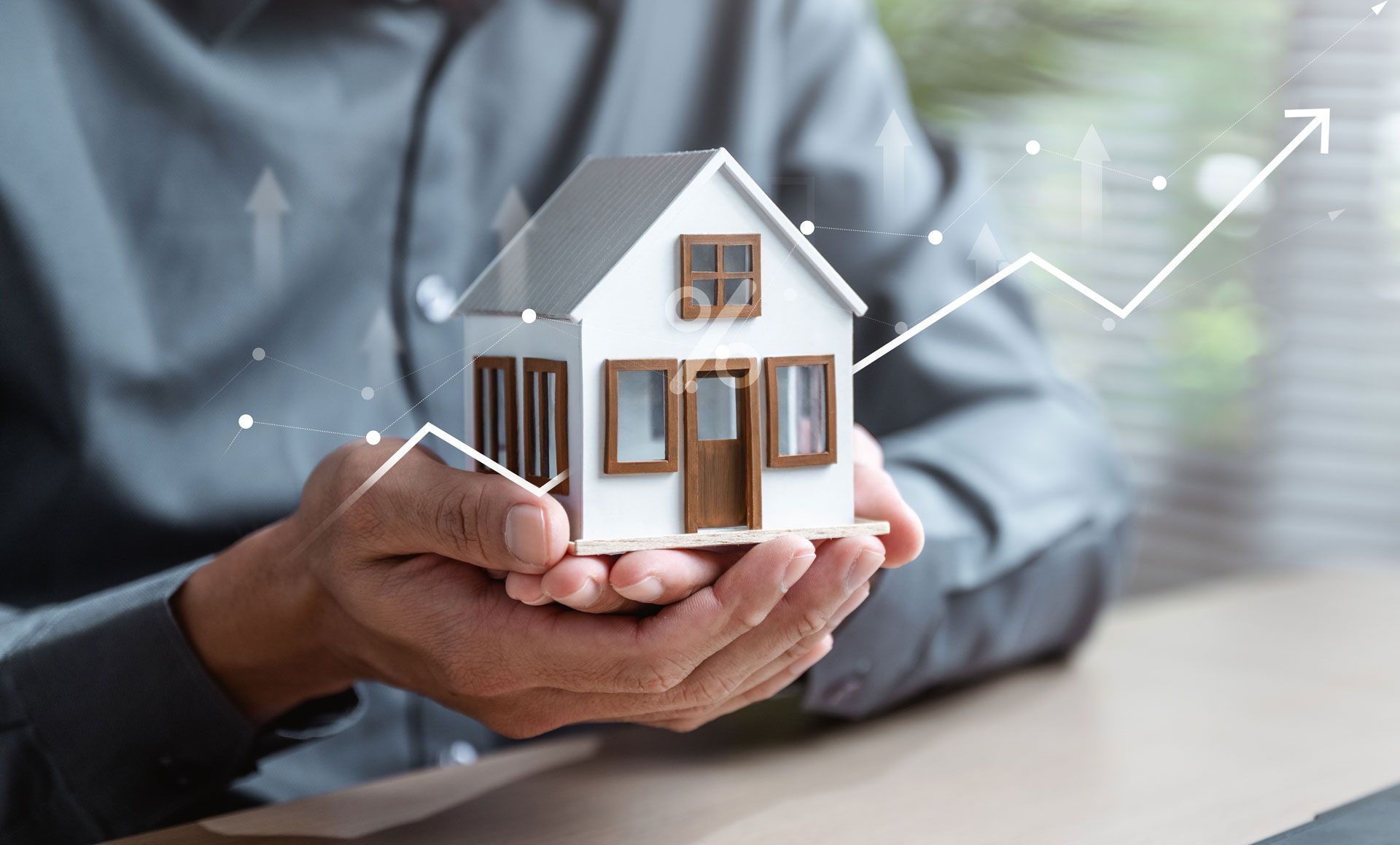 Hands holding a small white house model with a rising line graph in the background, suggesting real estate growth