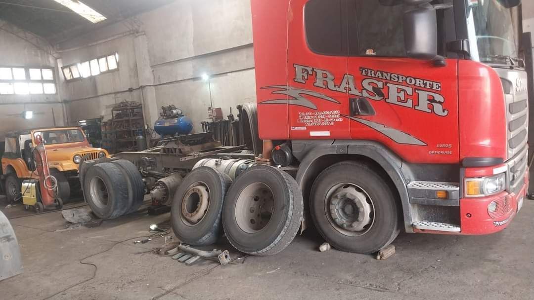 Un camión semirremolque rojo en un garaje con las ruedas sueltas y un mecánico trabajando al fondo.