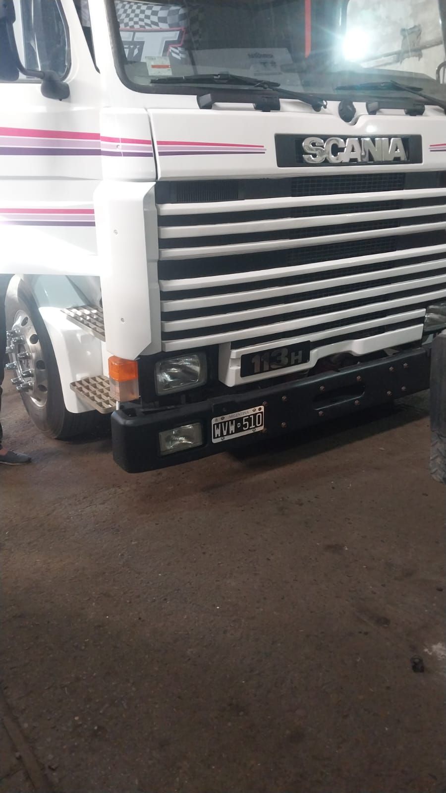 Vista frontal de un camión Scania blanco con franjas rosas estacionado en el interior.