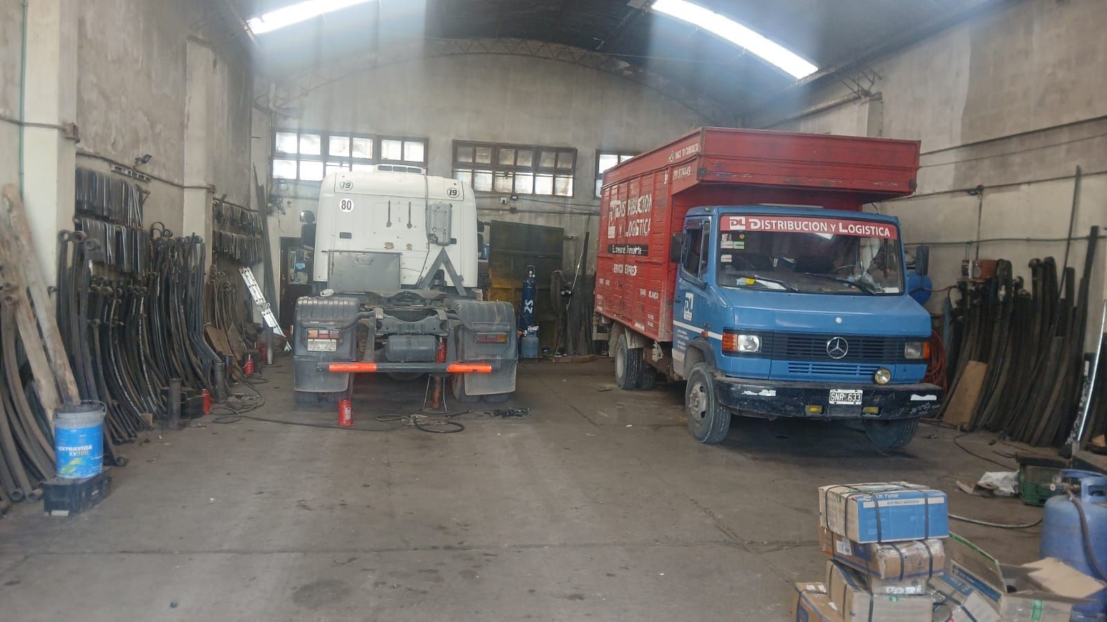 Taller mecánico con un camión azul y un remolque en el interior, herramientas alineadas en las paredes.