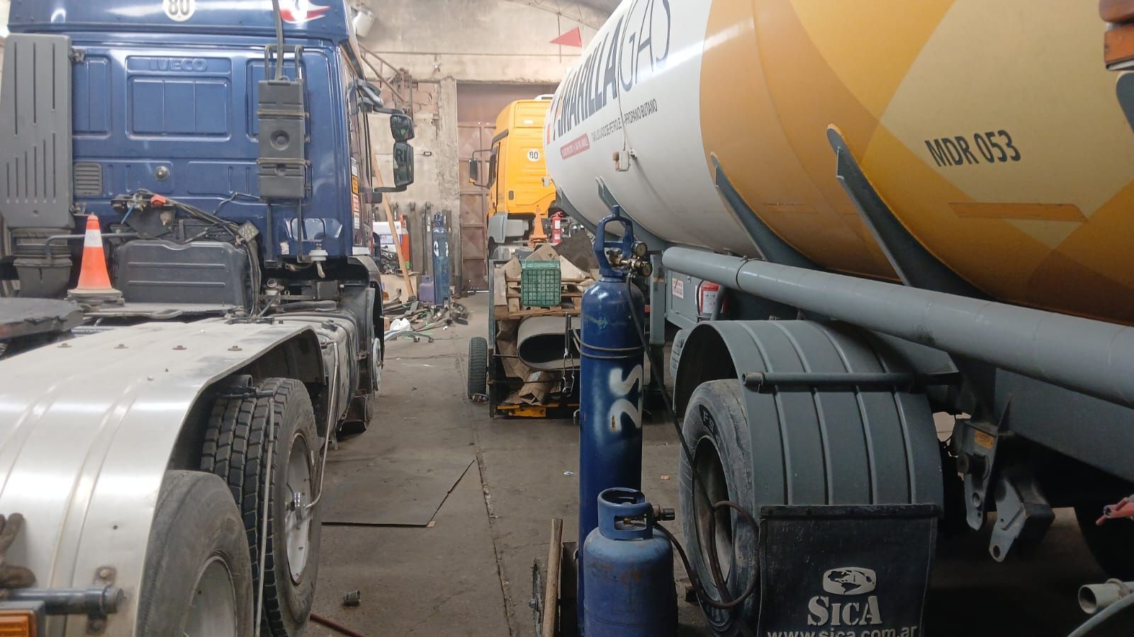 Camión hormigonera en un estrecho callejón industrial con maquinaria azul y conos de tráfico naranjas.