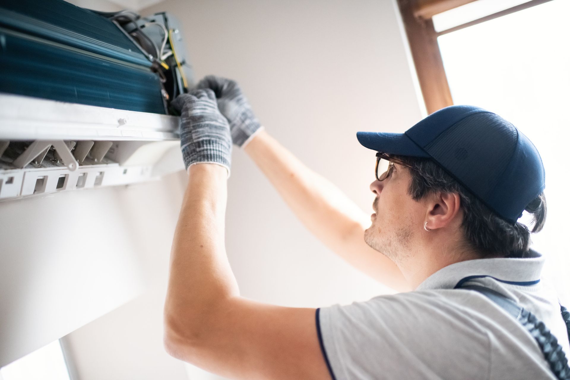 Technician in gloves inspects wall-mounted AC near bright window.