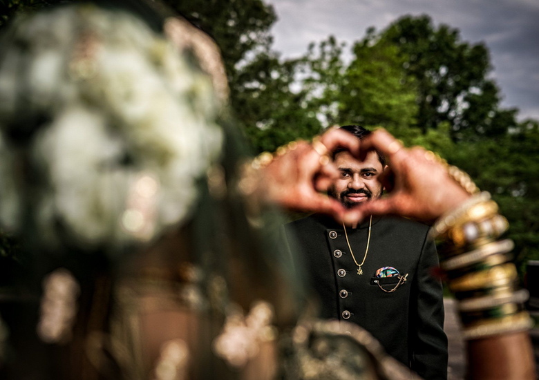 Smiling man forming a heart shape with his hands outdoors, with blurred flowers in the foreground and trees behind.