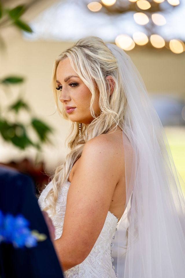 Blonde bride in a white veil and dress, looking over her shoulder at an outdoor wedding.