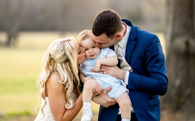 Two adults kissing a baby in a blue outfit outdoors, with a softly blurred background