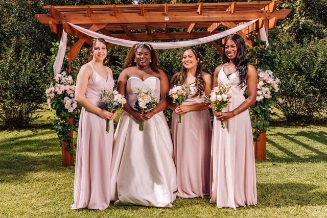 Four women in pink dresses holding bouquets under a wooden pergola in a garden