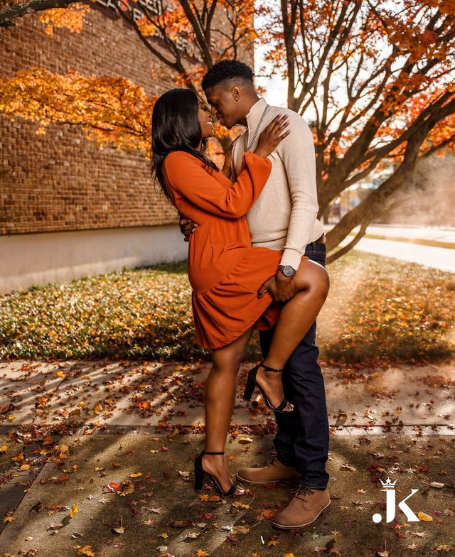 Couple embracing outdoors in autumn, with orange leaves and brick wall in the background.