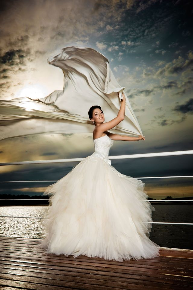 Woman in a white wedding gown twirling a long veil on a wooden dock at sunset