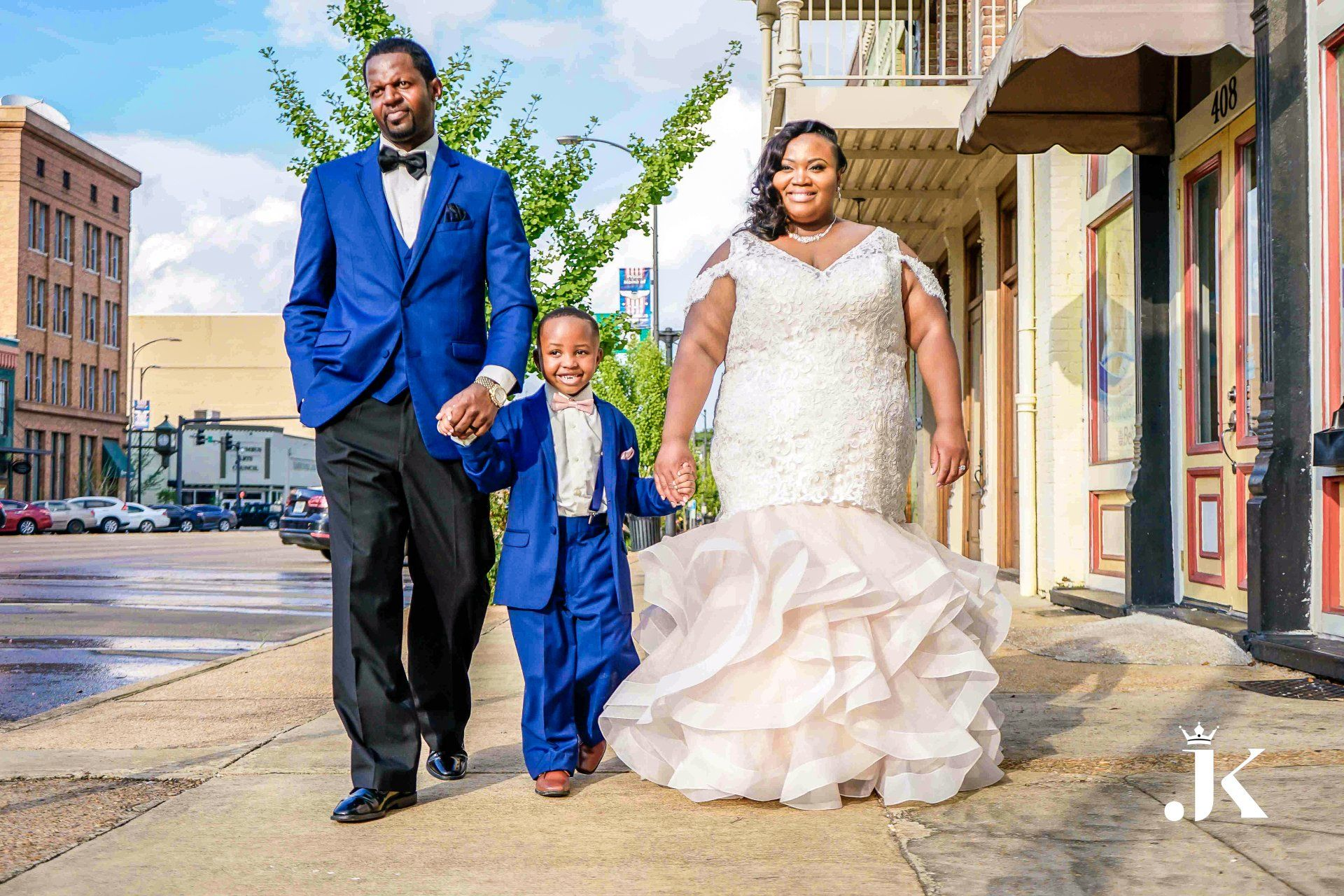 A couple in blue and white formal wear walk with a child on a sunny street.