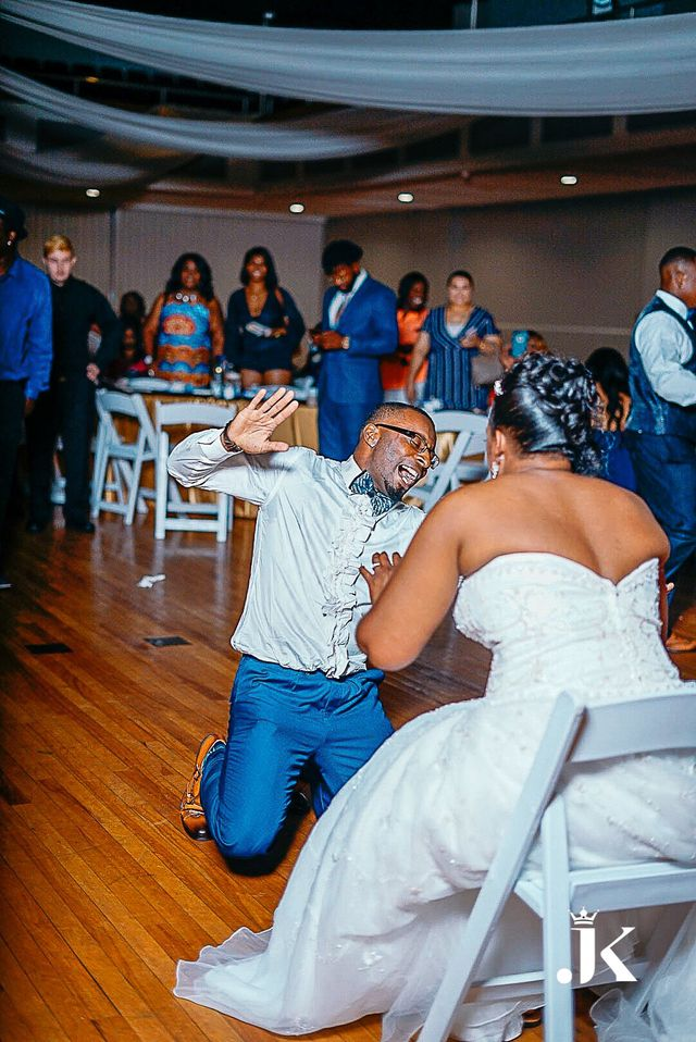Man kneels and proposes to a seated woman in a white dress at an indoor event hall.