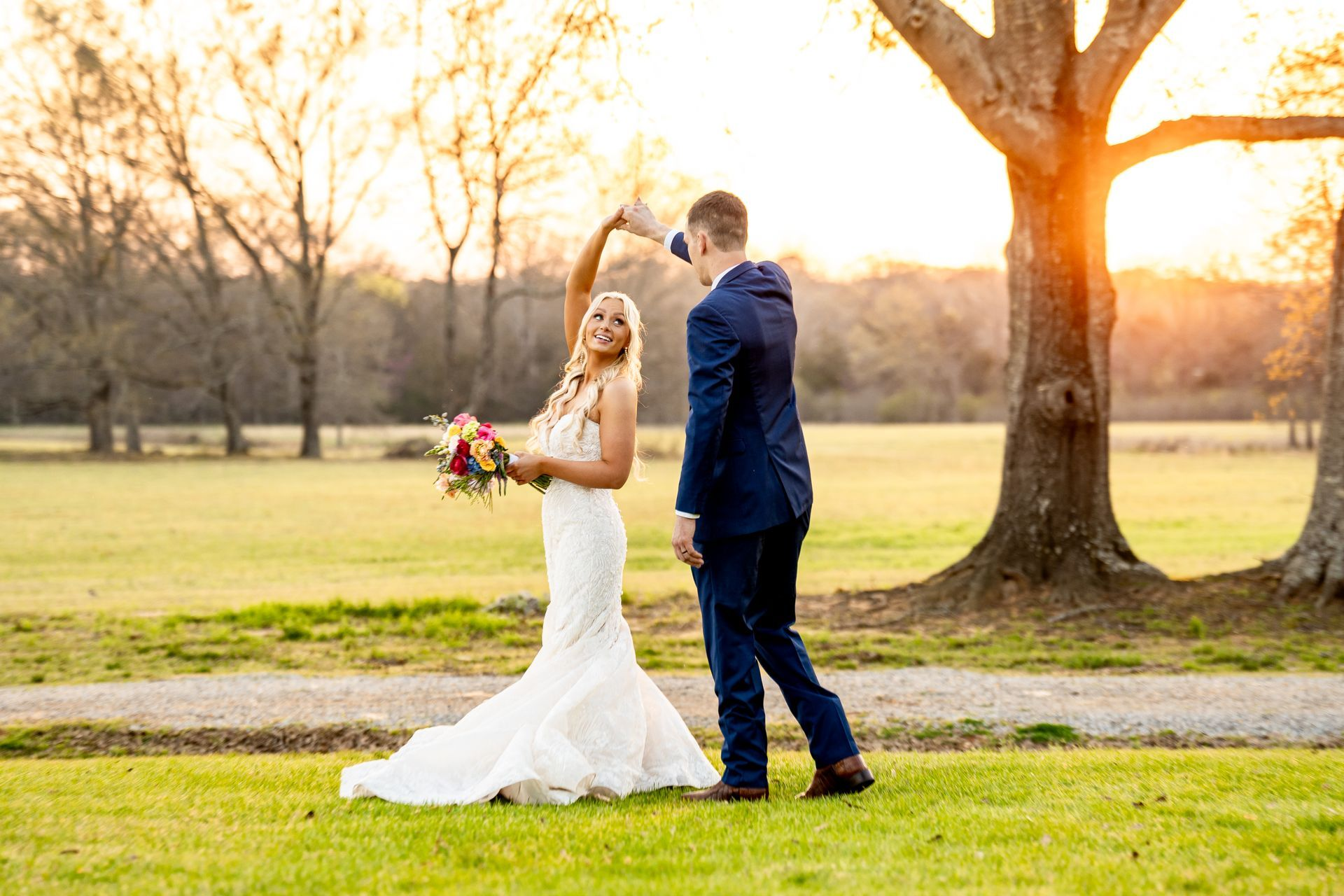 Bride and groom standing together outdoors at sunset, holding hands in a grassy park near a large tree
