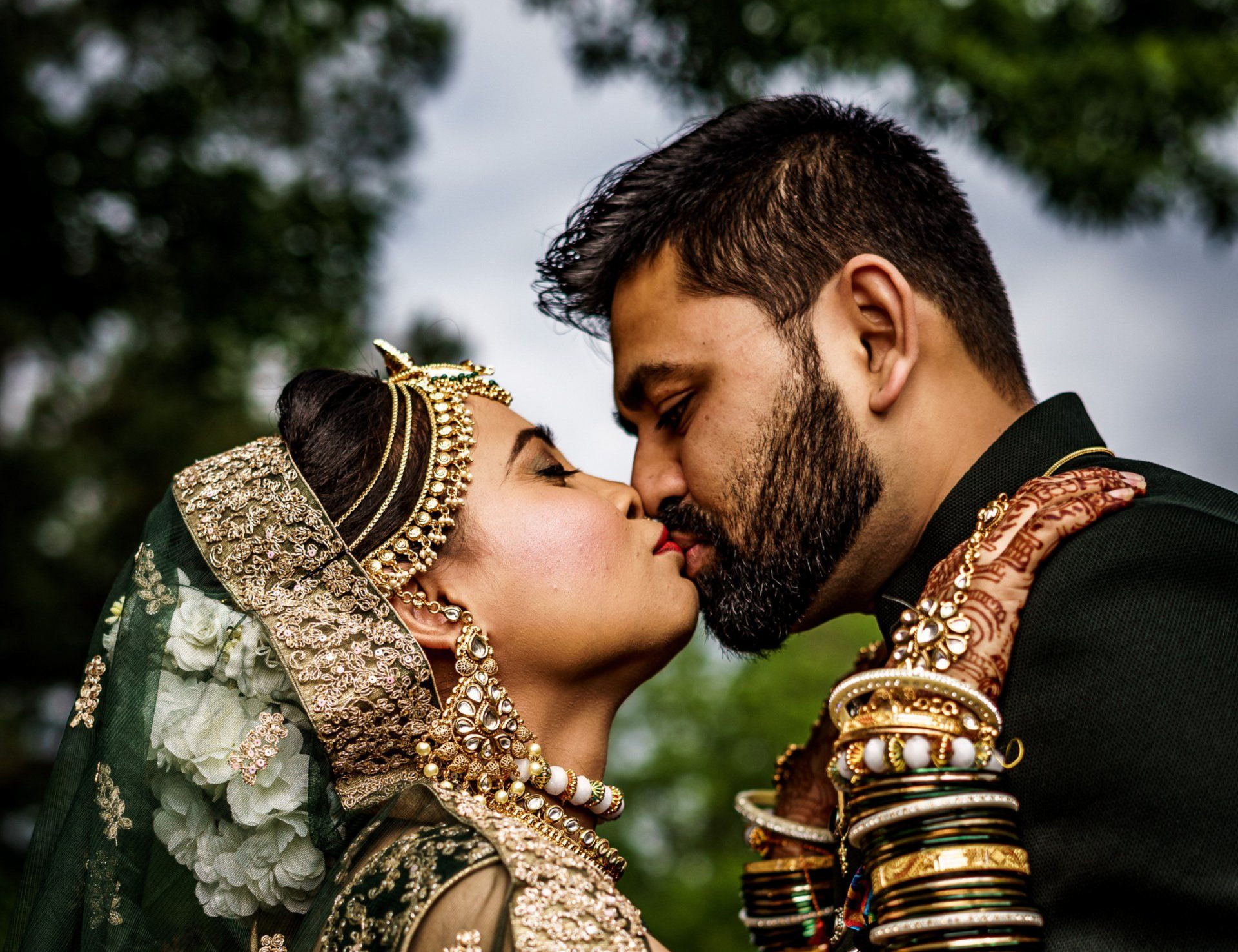 Bride and groom in ornate wedding attire sharing a kiss outdoors, with greenery in the background