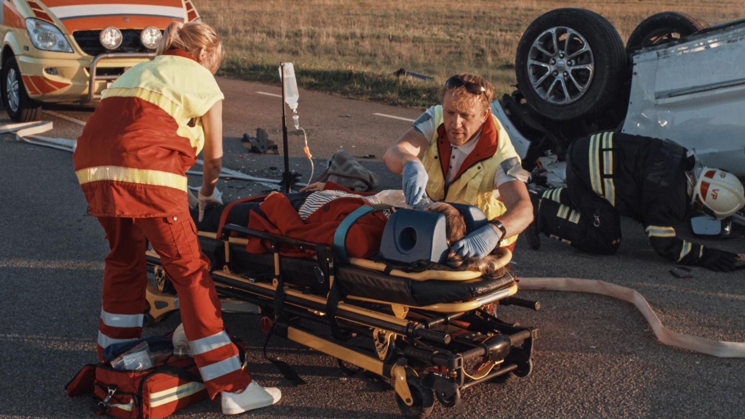 Emergency medical personnel treat a patient on a stretcher at a car accident scene; overturned vehicle visible.
