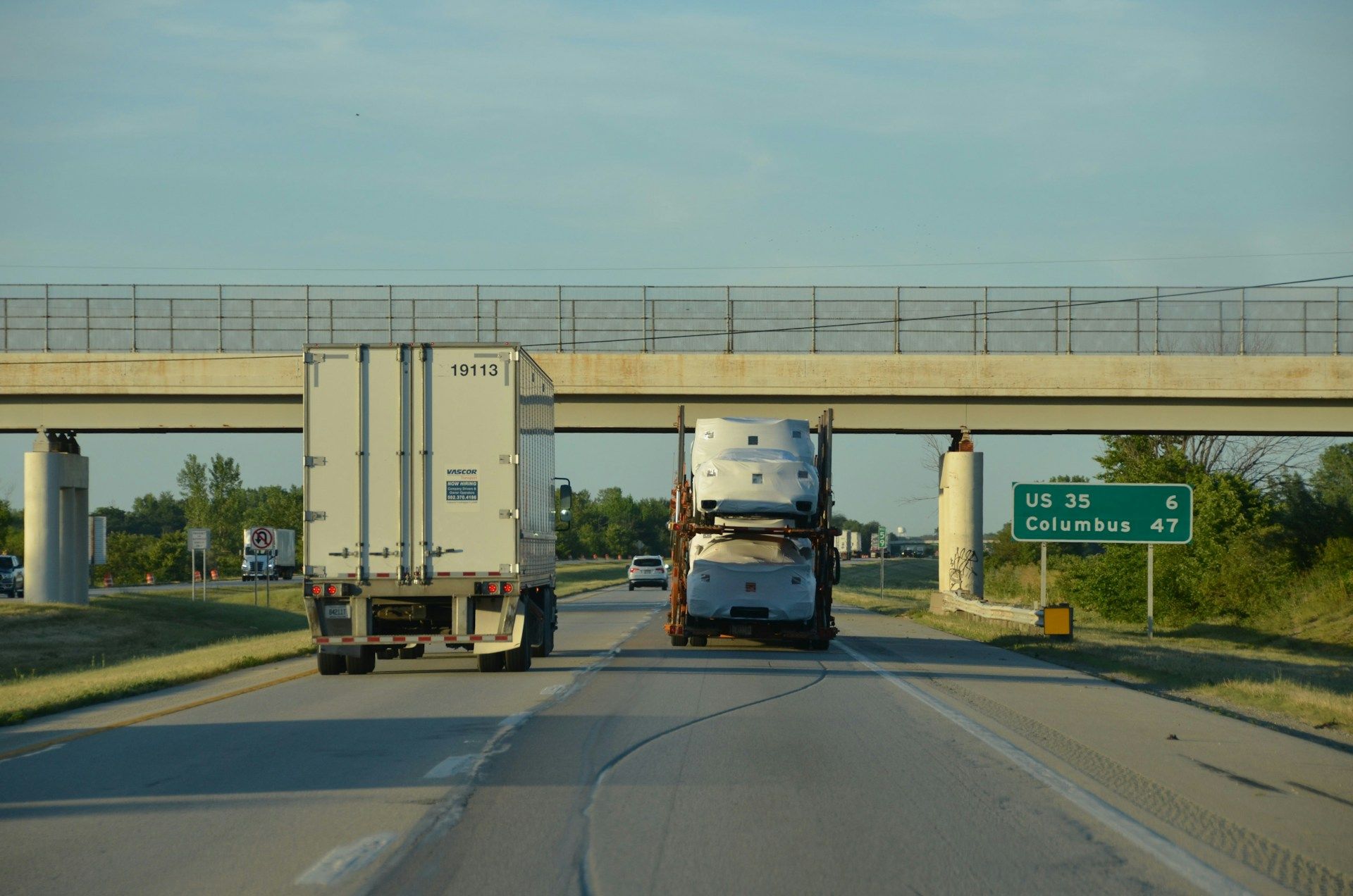 Two semi-trucks driving on highway under an overpass with a green road sign.