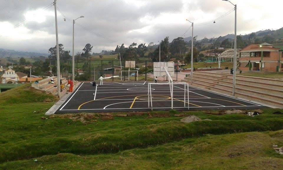 Cancha al aire libre con líneas pintadas y barandillas en una ladera, rodeada de césped, casas y cielo nublado.