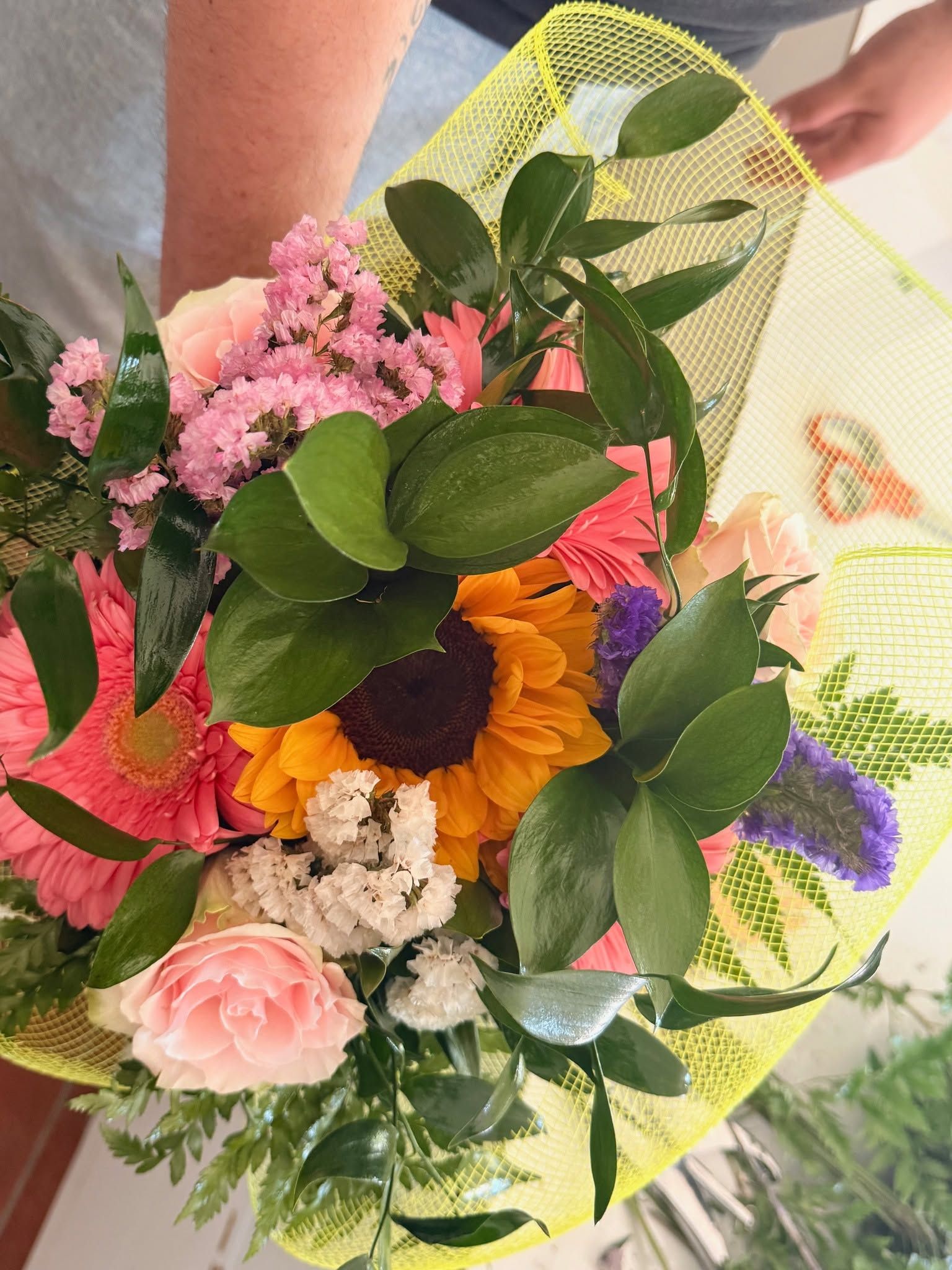Bouquet da tenere in mano, composto da fiori rosa, arancioni, viola e bianchi con foglie verdi, avvolto in una rete gialla.