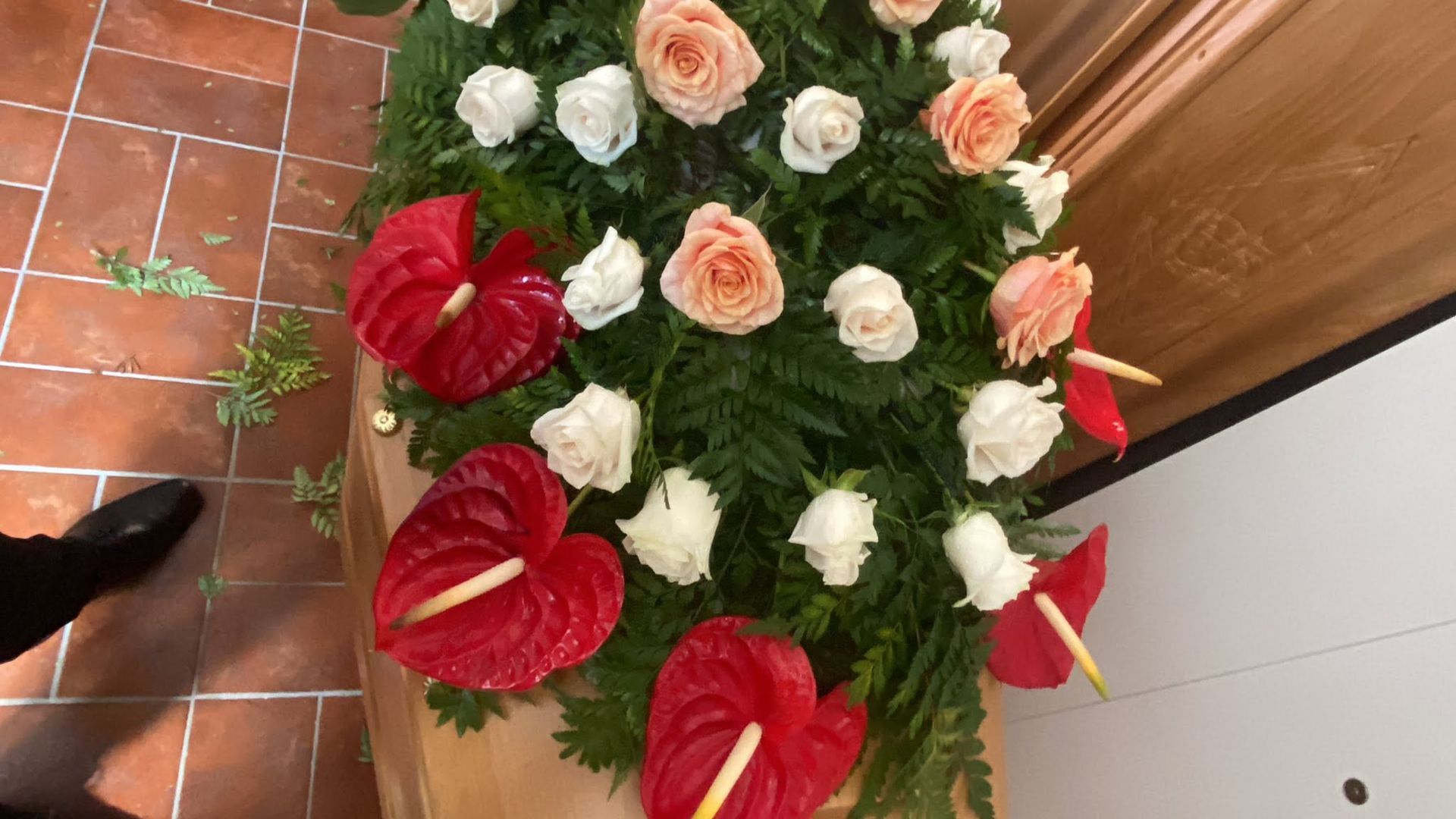 Composizione floreale rossa e bianca con rose e anthurium su un pavimento piastrellato vicino a una parete di legno.