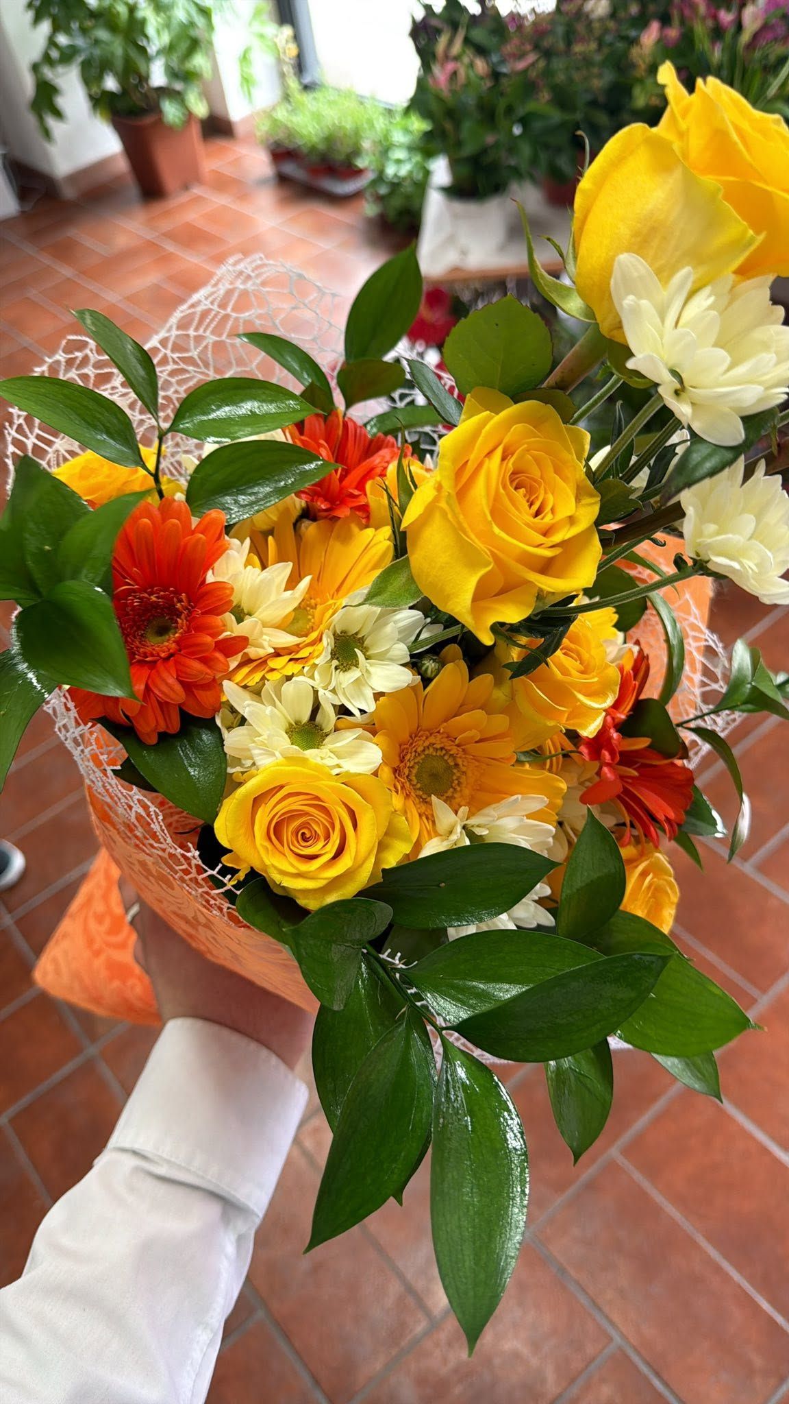 Bouquet di fiori gialli e arancioni con foglie verdi, tenuto a mano su un patio piastrellato.
