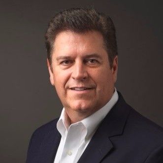Studio headshot of a man in a dark suit jacket and white shirt, smiling against a gray background