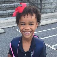 Girl with a red bow in her hair smiles, wearing a navy blue polo shirt and carrying a backpack outside.