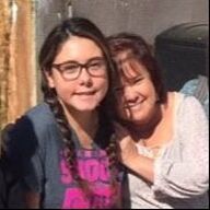 Woman and young person smiling close together outdoors; the young person wears glasses.