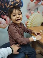 Young child smiling, seated on someone's lap, wearing brown hoodie, arm band, seated near table, indoors.