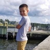 Boy standing on a dock, smiling, arms crossed. Wearing a light blue shirt and tan pants, waterside setting.