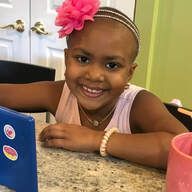 Smiling child with pink flower in hair, wearing jewelry, leaning on a table.