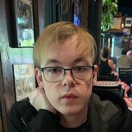 Person with glasses looking at the camera in a dimly lit setting.