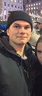 Man wearing a black beanie and jacket smiles, possibly outdoors, with lights in background.