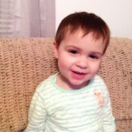 Toddler with short brown hair smiles while sitting on a textured couch.
