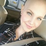 Woman with a shaved head smiles in a car, wearing a plaid shirt and seatbelt.