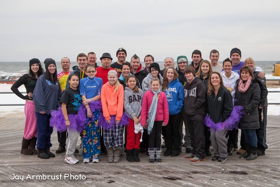 Group of people by a cold body of water, some wearing tutus, smiling. Overcast sky.