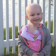 Young child with bald head, wearing a pink princess dress and gray jacket, smiles in front of a white picket fence.