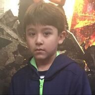 Boy in a blue hoodie, green trim, looking at the camera against a background of fire and rubble.
