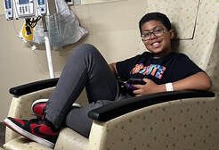 Boy in glasses and black shirt smiles, sits in a hospital chair, with IV stand in background.
