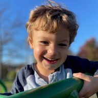 Smiling child with light brown hair, holding a green object outdoors on a sunny day.