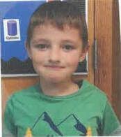 Boy in green shirt smiles in front of a classroom poster.
