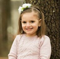 Girl with brown hair wearing a pink sweater and floral headband smiling. Leaning against a tree.