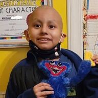 Child with a shaved head smiles, holding a blue monster toy. Wearing a mask, in front of a colorful wall.