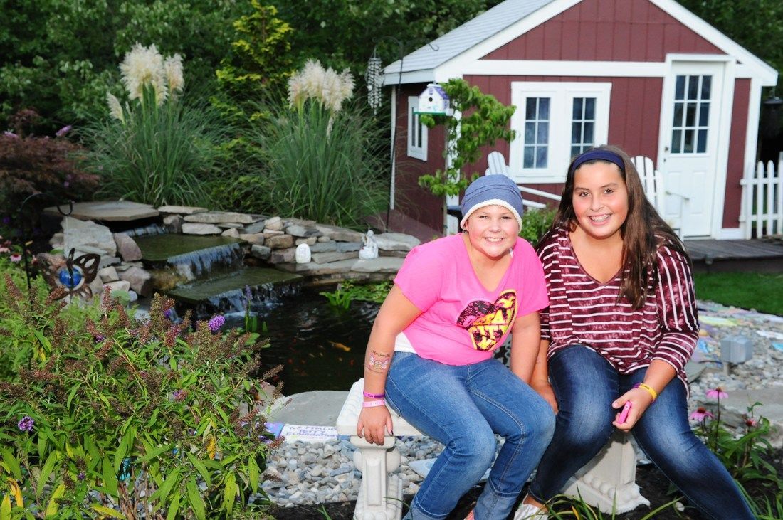 Two girls sit near a pond with a small red shed. One wears a pink shirt, the other a striped shirt. Lush greenery surrounds them.